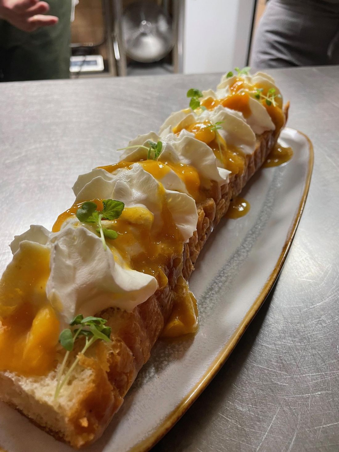 Un dessert sur un plat allongé : tranche dorée, crème, mangue et jeunes pousses